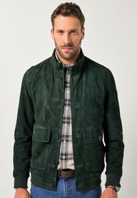 Giacca in suede verde con colletto rialzato, chiusura con bottoni e due tasche frontali. Indossata sopra una camicia a scacchi, con una texture liscia.
