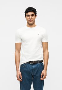 Άντρας με κοντά σγουρά μαλλιά που φοράει έναν απλό λευκό T-shirt και μπλε τζιν με μαύρη ζώνη, στέκεται μπροστά σε λευκό φόντο.