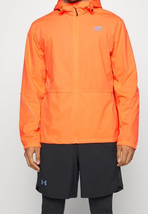 Fel oranje capuchonjack met voorrits, gedragen met zwarte sportshorts en leggings, geen zichtbaar gezicht of achtergronddetails.