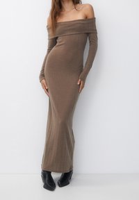 Vestido largo ajustado de un solo hombro en tela acanalada marrón, con mangas largas y corte inferior recto. Combinado con botas negras.