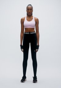 Brassière de sport rose avec un logo blanc, leggings noirs avec une ceinture "PRO NIKE", et chaussures de sport noires. Gants aux mains pour la prise.