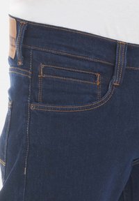Jeans de mezclilla azul oscuro con costuras en contraste, un parche de cuero en la cintura y un bolsillo trasero con un detalle mínimo.