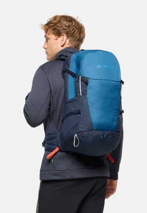 Homme blond portant une veste gris foncé et un pantalon noir, transportant un grand sac à dos de randonnée bleu et marine avec des sangles de taille.