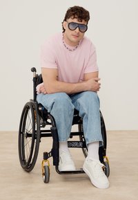 Joven con gafas de sol, camisa rosa, vaqueros azules y zapatillas blancas sentado en una silla de ruedas negra sobre un suelo claro y un fondo sencillo.