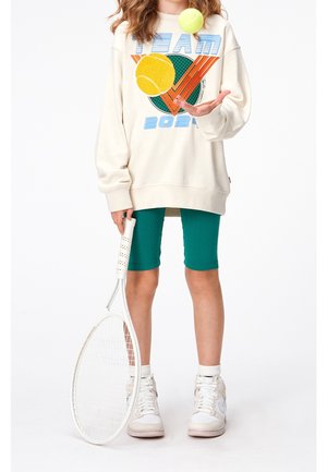 Enfant portant un sweat-shirt surdimensionné et un short bleu sarcelle, tenant une raquette de tennis blanche et lançant deux balles de tennis devant un fond blanc.