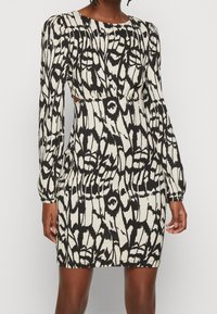 Robe à manches longues noire et blanche à motifs, avec une silhouette ajustée, un tissu texturé, un col rond et des découpes à la taille.