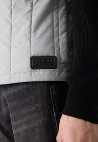 Casaco acolchoado cinza-claro com mangas em malha preta e um patch com o logótipo da Barbour International na parte inferior esquerda.