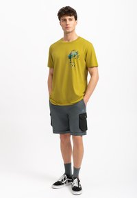 Olijfgroen t-shirt met abstracte grafische print, gecombineerd met grijze cargoshorts met zwarte zijzakken. Het model draagt zwarte sneakers.