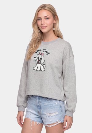 Giovane donna con lunghi capelli biondi che indossa una felpa grigia con un cane dei cartoni animati e il testo "Love Dog", abbinata a pantaloncini di jeans chiari strappati.