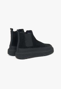 Black suede ankle boots with elastic side panels, thick black rubber soles, and textured detailing at the base. Minimalist design.