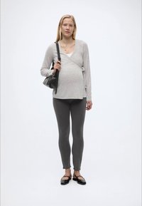 Έγκυος γυναίκα που φοράει ανοιχτό γκρι πουλόβερ τύπου wrap, σκούρο γκρι κολάν, μαύρα φλατ παπούτσια, κρατώντας μαύρη τσάντα ώμου μπροστά σε απλό λευκό φόντο.