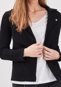 Donna che indossa un blazer nera testurizzata sopra una t-shirt bianca, mentre aggiusta la parte anteriore del blazer con entrambe le mani, senza viso in mostra.