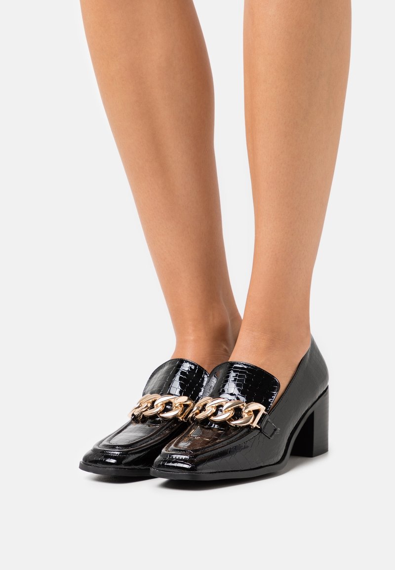 Mocassins à talons en cuir noir texturé crocodile avec un grand détail chaîne en or, portés sur des jambes nues sur un fond clair uni.