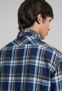 Camisa a cuadros en tonos azul, marrón y blanco. Cuello con botones y tela texturizada con costuras en contraste. Vista trasera.
