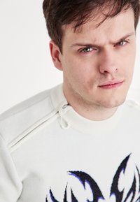 Λευκό πουλόβερ με σχέδιο φλόγας σε μαύρο και μπλε, ριγέ γιακά και λεπτομέρεια πλαϊκού φερμουάρ. Λείο υλικό και σύγχρονος σχεδιασμός.