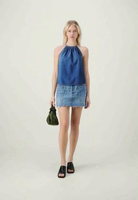 Haut en denim bleu à bretelles, associé à une jupe en denim bleu clair avec des bords effilochés et des coutures blanches. Sandales noires à bout ouvert et un sac vert.