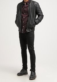 Giubbotto bomber in pelle nera sopra una camicia in plaid bordeaux, abbinato a jeans neri e scarpe nere con lacci e una texture liscia.