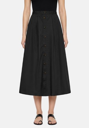 Donna che indossa una gonna midi nera a vita alta con bottoni frontali e pieghe, abbinata a un top nero senza maniche e sandali neri.