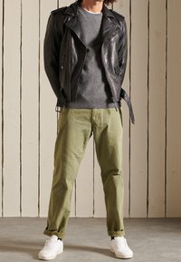 Veste de moto en cuir noir portée sur un pull en maille gris, associée à un pantalon vert olive et des baskets blanches. Matériaux texturés avec couture visible.