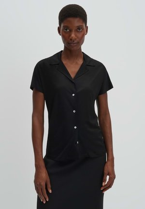 Frau trägt ein kurzärmeliges, schwarzes Hemd mit Kragen und Knöpfen, steht vor einem einfarbigen hellgrauen Hintergrund.