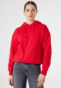 Sudadera con capucha roja, hecha de una mezcla de algodón, con un diseño recortado, puños acanalados y herrajes de cordón en tono dorado. Combinada con jeans grises.