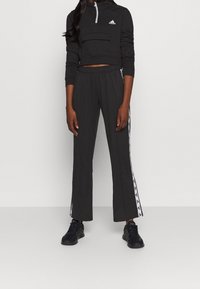 Černá krátká mikina s kapucí, přední kapsou a zipem, kombinovaná s černými tepláky s bílými pruhy po stranách a volným střihem. Černé tenisky.