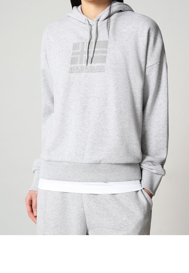 Persoon draagt een lichtgrijze capuchontrui en bijpassende joggingbroek, met één hand in de zak, effen achtergrond.