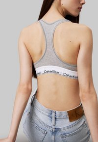 Sostenedor deportivo de algodón gris con una ancha banda blanca con el logo "Calvin Klein", visto desde la parte de atrás. Jeans de mezclilla azul claro parcialmente visibles.