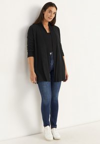 Frau in dunklem offenen Cardigan, schwarzem Top, blauen Skinny Jeans und weißen Sneakers steht an einer weißen Wand und blickt leicht zur Seite.