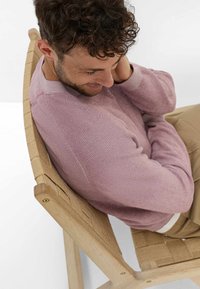 Een houten stoel met geweven beige stof. De zitting is schuin; de rugleuning heeft een kruispatroon. De persoon draagt een lichtroze trui.