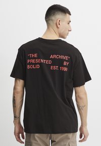 Juodas medvilninis marškinėliai su trumpomis rankovėmis, su raudonu tekstu ant nugaros: „ARCHYVAS“ „PATEIKTA SOLID EST. 1999.“
