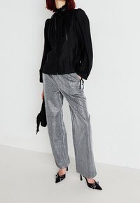 Blouse noire à manches longues avec volants et boutons, associée à un pantalon large à rayures argentées brillantes et des escarpins noirs à bout pointu ornés d'accents.