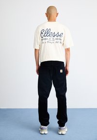 Kreminės spalvos trumpomis rankovėmis marškinėliai su mėlynu "Ellesse" logotipu, derinami su juodais veliūriniais pantalonais. Išskirtinis siuvinėjimas ir mažas logotipas ant galinio kišenės.