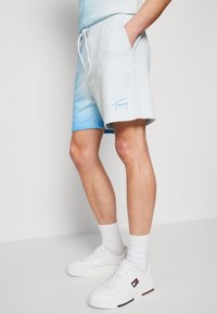 Světlé modré sportovní šortky s hladkým povrchem, vybavené bočními kapsami a stahovacími šňůrkami v pase. Na nohavici je vytištěno logo "Tommy" modrou barvou.