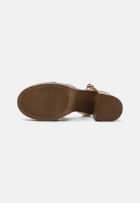 Semelle marron d'une sandale féminine avec un talon bas, semelle à motif étoile et boucle de sangle à la cheville, photographiée sur un fond blanc.