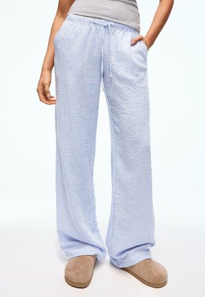 Personne portant un pantalon large rayé bleu clair et blanc avec les mains dans les poches et des chaussures beige à enfiler sur un fond uni.