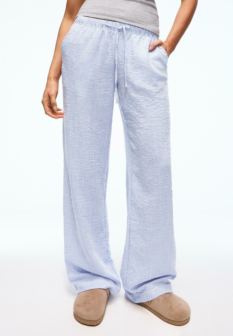 Personne portant un pantalon large rayé bleu clair et blanc avec les mains dans les poches et des chaussures beige à enfiler sur un fond uni.