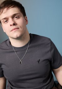 Γκρι μπλουζάκι με μικρό λογότυπο μαύρου πουλιού στο αριστερό στήθος, σε συνδυασμό με ασημένια αλυσίδα που φέρει ορθογώνιο κρεμαστό.