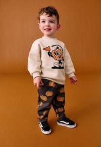 Sweat-shirt crème avec motif imprimé de citrouille cartoon, accompagné d'un pantalon sombre orné de motifs de citrouilles orange, et de baskets noires avec semelles blanches.