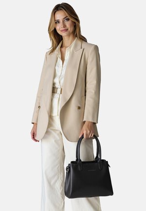 Mujer con blazer beige, camisa blanca y pantalones, sosteniendo un bolso de cuero negro con doble asa sobre un fondo liso.