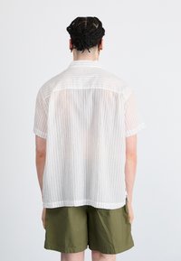 Kortermet, hvitstripet skjorte laget av lett materiale, sammen med mørkegrønne shorts. Skjorten har en avslappet passform og sømmet nederkant.