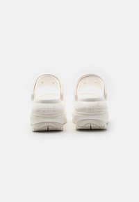 Crocs blancs avec une sangle arrière, présentant un matériau lisse et brillant, une forme arrondie et des semelles texturées pour une meilleure adhérence.