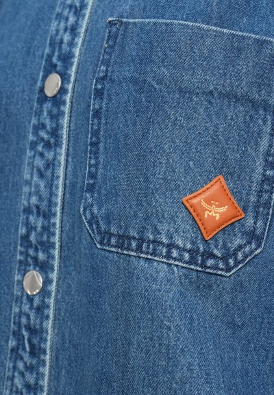 Cămașă din denim cu țesătură albastră, capse argintii și o buzunar cu etichetă din piele maro cu design embosat.