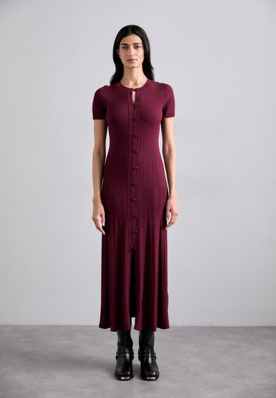 Robe tricotée côtelée bordeaux à manches courtes, devant à boutons, et fente haute sur la cuisse, associée à des bottines noires.