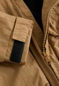 Giacca tan con una texture di tessuto morbido, chiusura a zip con un tirante unico e una pattina in Velcro sui polsini per la regolazione.