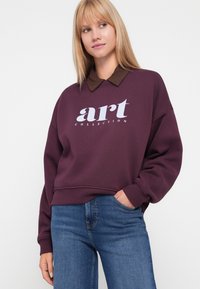 Felpa bordeaux con scollo rotondo, vestibilità over, e sottogiacca color marrone con colletto. Presenta la scritta bianca "art COLLECTION" sulla parte anteriore.
