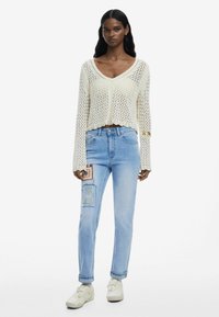Cremfarbener Strickpullover mit V-Ausschnitt, langen Ärmeln und gewelltem Saum. Hellblaue Jeans mit Patchwork-Details und weißen Sneakers.