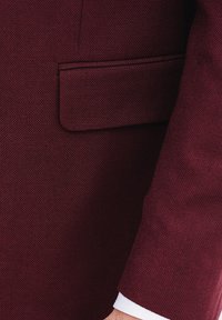 Bordowy blezer z teksturowanej tkaniny, z jedną kieszenią na piersi i kontrastowym ściegem. Rękaw kończy się na nadgarstku, odsłaniając mankiet białej koszuli.