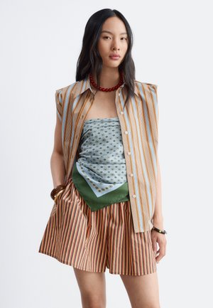 Femme aux longs cheveux noirs portant un débardeur rayé, un haut foulard à motif, un short rayé, un collier rouge et des bracelets sur fond blanc.