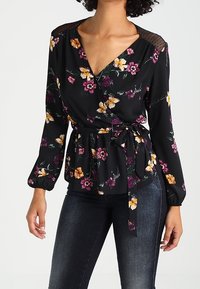 Blouse portefeuille noire avec motifs floraux en violet et jaune. Dotée de manches longues, d'une ceinture à nouer et d'un détail en maille sur les épaules.